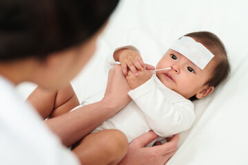 Obraz na płótnie Canvas mother using cotton bud to clean nose of sick baby with cool fever jel pad on bed