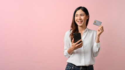 A woman holding smartphone and credit card on pink isolated background.