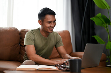 Happy Asian man using laptop on a sofa in the living room. working, searching online