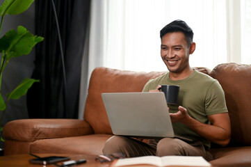 Happy millennial Asian man sipping coffee and using his laptop on a sofa in the living room