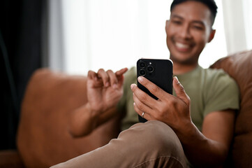 Happy Asian man using his smartphone and relaxing on a sofa in his living room.
