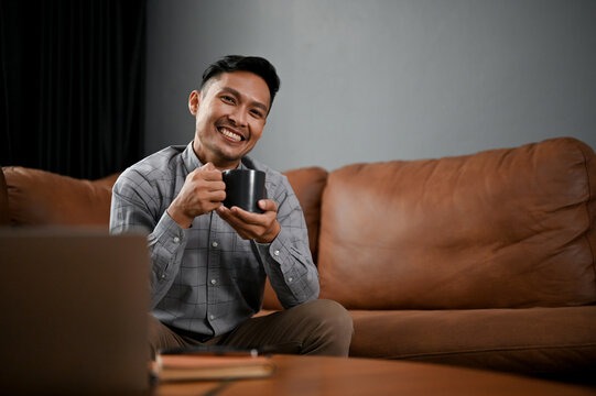 Smiling Asian man sitting on brown leather sofa with a coffee cup, relaxing on his free time