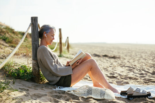 Mature woman wearing wireless in-ear headphones reading book at beach