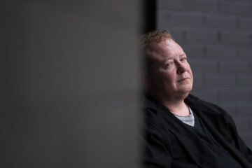 Although his powerchair is not visible, a disabled transman sits in his chair while posing for a portrait. He is surrounded by brick walls painted black. He has a calm and confident expression.