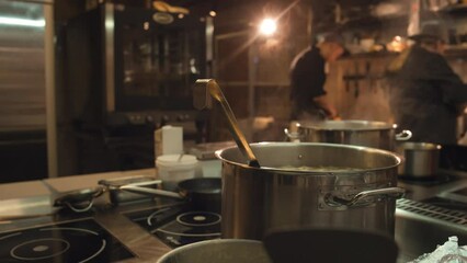 Medium close-up shot of steel pot of hot steaming soup cooking on induction stove in restaurant kitchen, and unrecognizable chefs working in background - Powered by Adobe