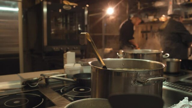 Medium close-up shot of steel pot of hot steaming soup cooking on induction stove in restaurant kitchen, and unrecognizable chefs working in background