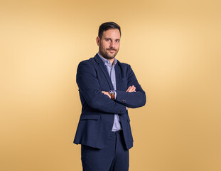 Obraz na płótnie Canvas Portrait of male manager with arms crossed posing confidently while standing isolated over beige background. Young businessman dressed in elegant blue suit looking at camera
