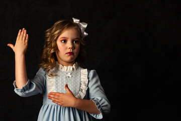 Adorable little doll cover girl gesturing hands at black background, looking away. Cute kid in blue dress and white bow on head isolated with light and shadow. Fashion image concept. Copy text space