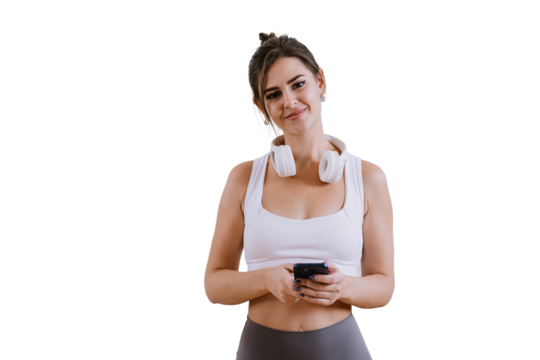 Attractive brunette American girl in sportswear and headphones on neck holds phone looks at camera smiles standing against transparent background.Sport, fitness. Young woman at dance class. Health. - Powered by Adobe