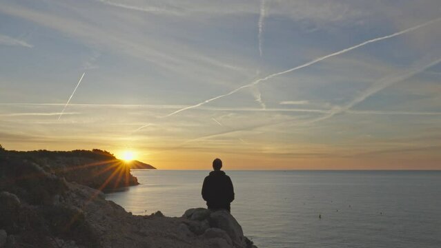 Timelapse captures man sitting on rocks experiencing beautiful golden sunrise by the seaside in Sitges, Spain