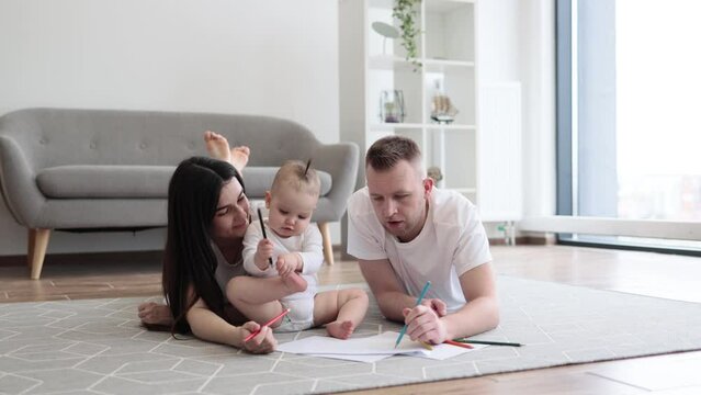Adorable little baby girl in white cozy bodysuit holding red pencil while sitting on floor with mindful mother and father on both sides. Adult people and kid enjoying first scribbles on paper at home.