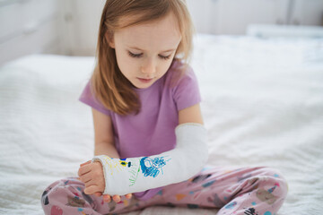 Adorable preschooler girl with a broken arm at home on the bed draws with felt-tip pens on an...