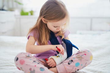Adorable preschooler girl with a broken arm at home on the bed draws with felt-tip pens on an...