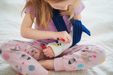 Adorable preschooler girl with a broken arm at home on the bed draws with felt-tip pens on an...