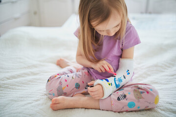 Adorable preschooler girl with a broken arm at home on the bed draws with felt-tip pens on an...