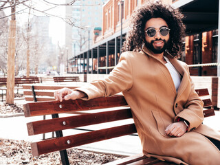 Handsome smiling hipster model.  Unshaven Arabian man dressed in brown coat clothes. Fashion male with long curly hairstyle posing outdoors in street. Cheerful and happy. In sunglasses. At sunny day