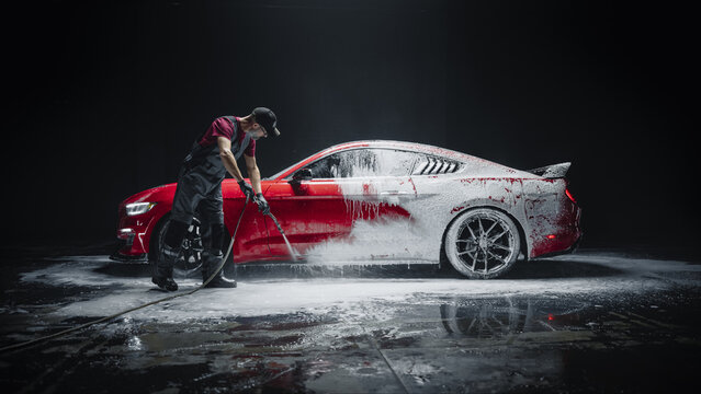 Car Wash Specialist Using Pressure Washer to Rinse a Red Modern Sportscar. Adult Man Washing Away Dirt, Preparing a Tuned Car for Detailing. Creative Cinematic Photo in Studio