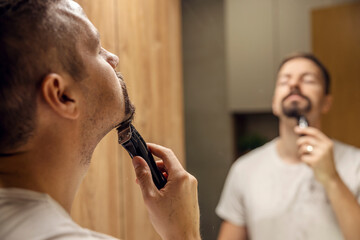 Close up of a bearded man shaving his beard with shaver in front of the mirror in bathroom.