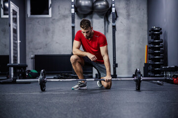 Naklejka na ściany i meble Preparing for strong muscle-burning training. A young attractive man in red and grey sportswear sets up barbell weights in the gym. Fit young man looking focused on practice, sports discipline
