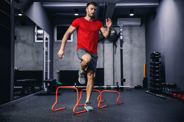 Fototapeta na wymiar Lateral skipping of small orange obstacles and performing warm-up exercises. A photo of the whole body of a handsome man in the dark atmosphere of a gym. Individual training with hurdles