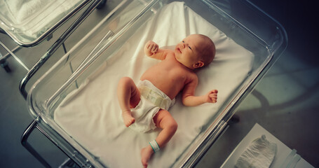 Adorable Small Caucasian Newborn Child Lying in Hospital Bed in a Nursery Clinic. Little Playful and Healthy Baby. Medical Health Care, Maternity and Parenthood Concept. High Angle Close Up Portrait - obrazy, fototapety, plakaty