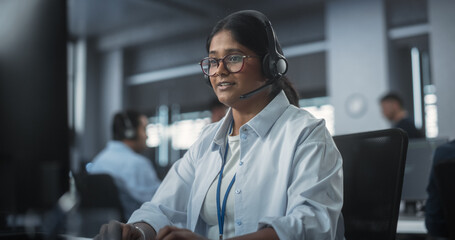 Portrait of a Smart Young Indian Specialist Working in a Technological Startup Help Desk Department, Using Computer in a Diverse Office. Female Wearing Glasses and Headphones with a Microphone
