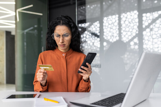 Upset and disappointed hispanic businesswoman inside office at workplace, woman holding bank credit card and phone, employee cheated trying to make online purchase, money transfer error.