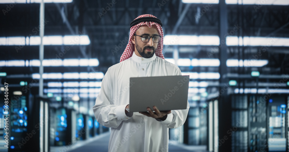 Poster Portrait of a Middle Eastern Data Center Administrator Wearing Traditional White Clothes, Using Laptop Computer in a Modern Technological Server Farm Facility. Arab Cybersecurity Expert at Work