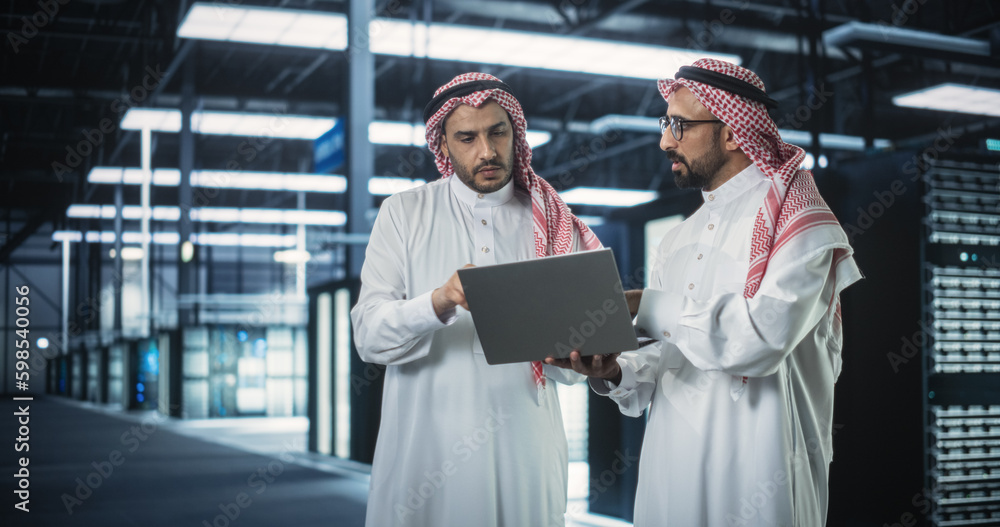Aufkleber Middle Eastern Computer Engineer and Cybersecurity Professional Work in Server Room, Talk, Perform System Maintenance Check with Laptop Computer. Specialists Doing Database Diagnostics in Data Center