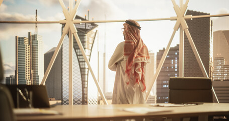Successful Muslim Businessman in Traditional White Outfit Standing in His Modern Office Looking out of the Window on Big City with Skyscrapers. Successful Saudi, Emirati, Arab Businessman Concept