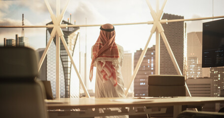 Successful Muslim Businessman in Traditional White Outfit Standing in His Modern Office Looking out of the Window on Big City with Skyscrapers. Successful Saudi, Emirati, Arab Businessman Concept