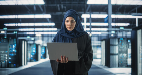 Muslim Female Data Center IT Engineer Standing in a Room with Server Racks. Cloud Computing Architect Uses Laptop Computer for Servicing the System in Cyber Security and Data Protection Facility