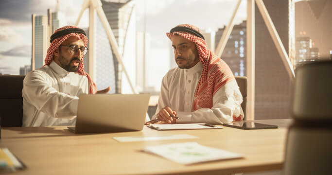 Two Young Muslim Financial Advisers Use Laptop Computer to Discuss a Stock Market Strategy in a Modern Industrial Office. Arab Managers Work in a Banking Research and Development Center