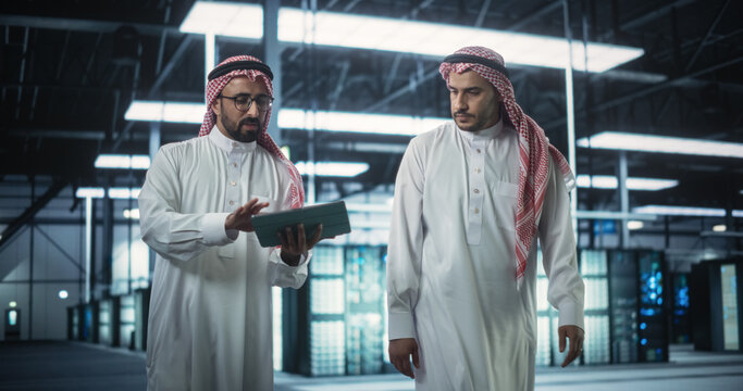 Middle Eastern Computer Engineer and Cybersecurity Specialist Work in Server Room, Talk, Perform System Maintenance Check with Laptop Computer. Specialists Doing Database Diagnostics in Data Center