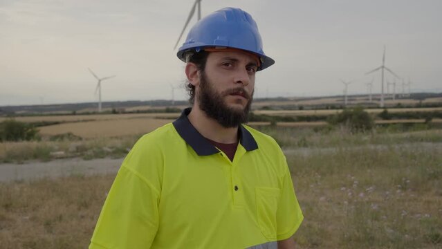 Young Engineer at Natural Energy Wind Turbine facility to inspect operation of large wind turbines electrical power. Male technician poses for photo looking at camera serious