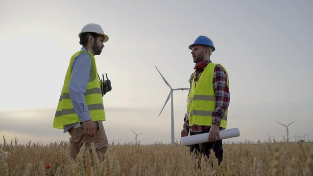 Engineer and technician shaking hands to implement new sustainable project in agricultural field wind turbines. Professional technicians close deal outside wind farm. Clean renewable