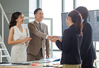Asian professional successful businesswoman standing smiling shaking hands greeting with unrecognizable businessman in formal business suit while male female colleagues clapping hands celebrating.