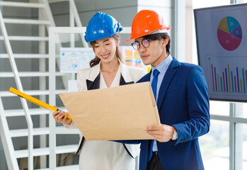 Asian professional successful female businesswoman and male businessman engineering employees in formal business suit and safety helmet standing holding blueprint and water level gauge discussing