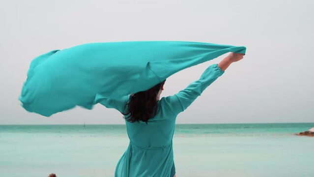 Woman in turquoise airy dress with pareo over head stands on ocean beach in windy day admires seascape. Brunette female enjoying summer holidays vacations on tropical resort. Travel, Generative AI