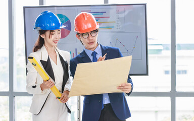 Asian professional successful female businesswoman and male businessman engineering employees in formal business suit and safety helmet standing holding blueprint and water level gauge discussing