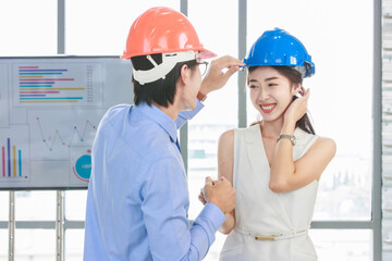 Asian professional successful male businessman holding water level gauge helping adjusting female businesswoman engineering safety hardhat standing smiling with document clipboard in meeting room