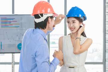 Asian professional successful male businessman holding water level gauge helping adjusting female businesswoman engineering safety hardhat standing smiling with document clipboard in meeting room