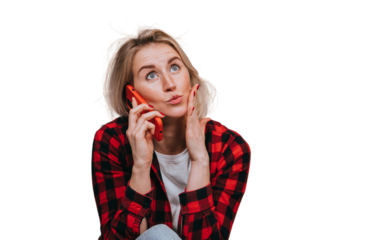 Excited blond young woman in plaid shirt talking by phone touches face with wide opened eyes sits home against transparent background with view on ocean and sunset. Amazed Swedish girl hearing gossip