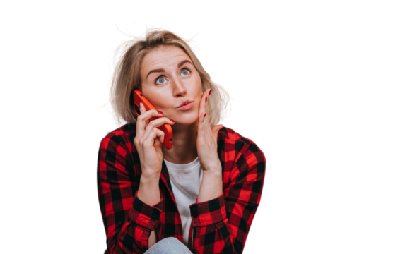 Excited blond young woman in plaid shirt talking by phone touches face with wide opened eyes sits home against transparent background with view on ocean and sunset. Amazed Swedish girl hearing gossip