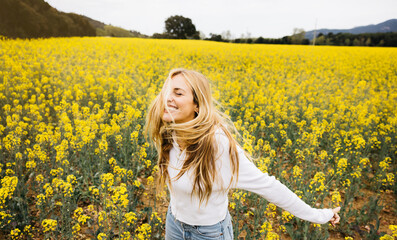 Naklejka na ściany i meble Beautiful young blonde cheerful woman, wearing a hat, poses amidst a field of blooming yellow rapeseed flowers