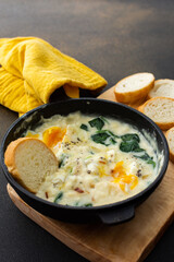Healthy breakfasts or dinners, fried eggs with cheese, spinach leaves and coconut milk in a cast-iron pan, sliced baguette on a cutting board on a brown background