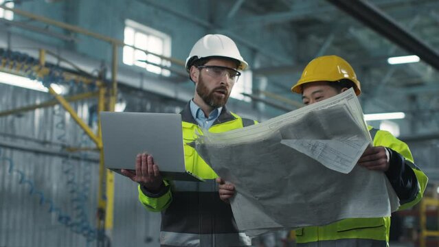 Architect in hard hat and protective glasses. Two workers are talking at construction site. Engineer holding laptop. Male explains something to the subcontractor and gestures.