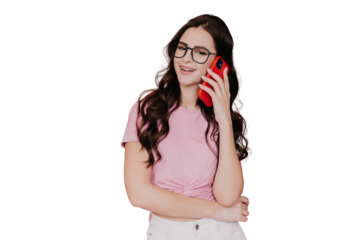 Beautiful brunette girl in glasses, pink t-shirt and white pants making call using phone over transparent background. Pretty young woman with wavy long hair looking at camera talking by cell phone.