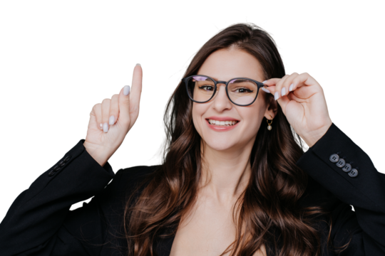 Gorgeous brunette young woman with loose wavy hair adjusts spectacles on face toothy smiles looking at camera points up by index finger at empty space on transparent background. Mockup