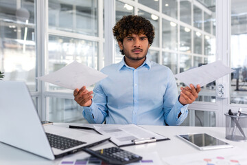 Portrait of angry disappointed financier inside office at workplace, hispanic businessman holding contracts reports in hands and looking nervously at camera, upset man behind paper work. - obrazy, fototapety, plakaty